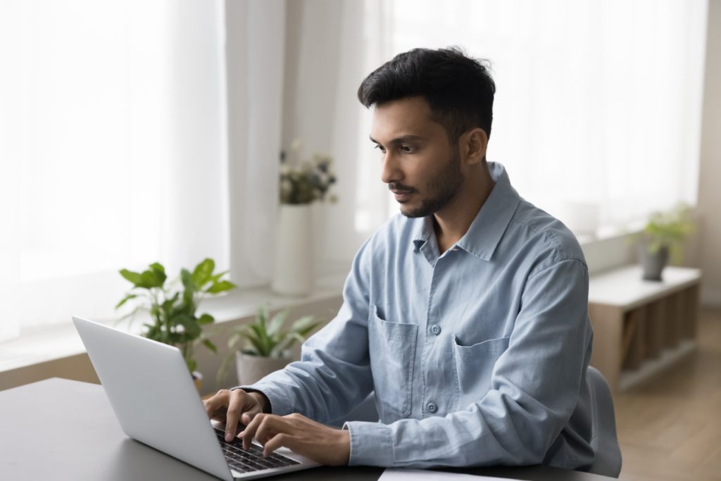 Focused young Indian professional man working on business project at laptop, typing, sitting at table in home office. Freelance manager using Internet technology, job application on computer